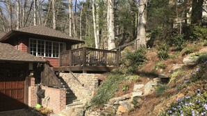 Une cabane avec un escalier en pierre menant à une terrasse en bois, le tout entouré d’une zone boisée.