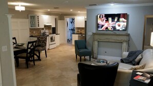 A living room with a fireplace, a dining area with chairs, a kitchen with cabinets, and a TV mounted on the wall. - Temporary living that feels like home where you can relax and be yourself. (Maitland)