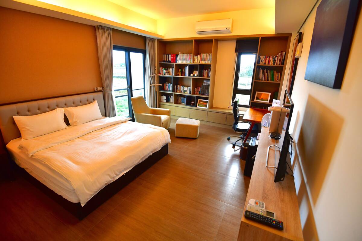 Une chambre d’hôtel avec un grand lit, un bureau équipé d’un ordinateur et d’une télévision, une bibliothèque et une fenêtre avec des rideaux.
