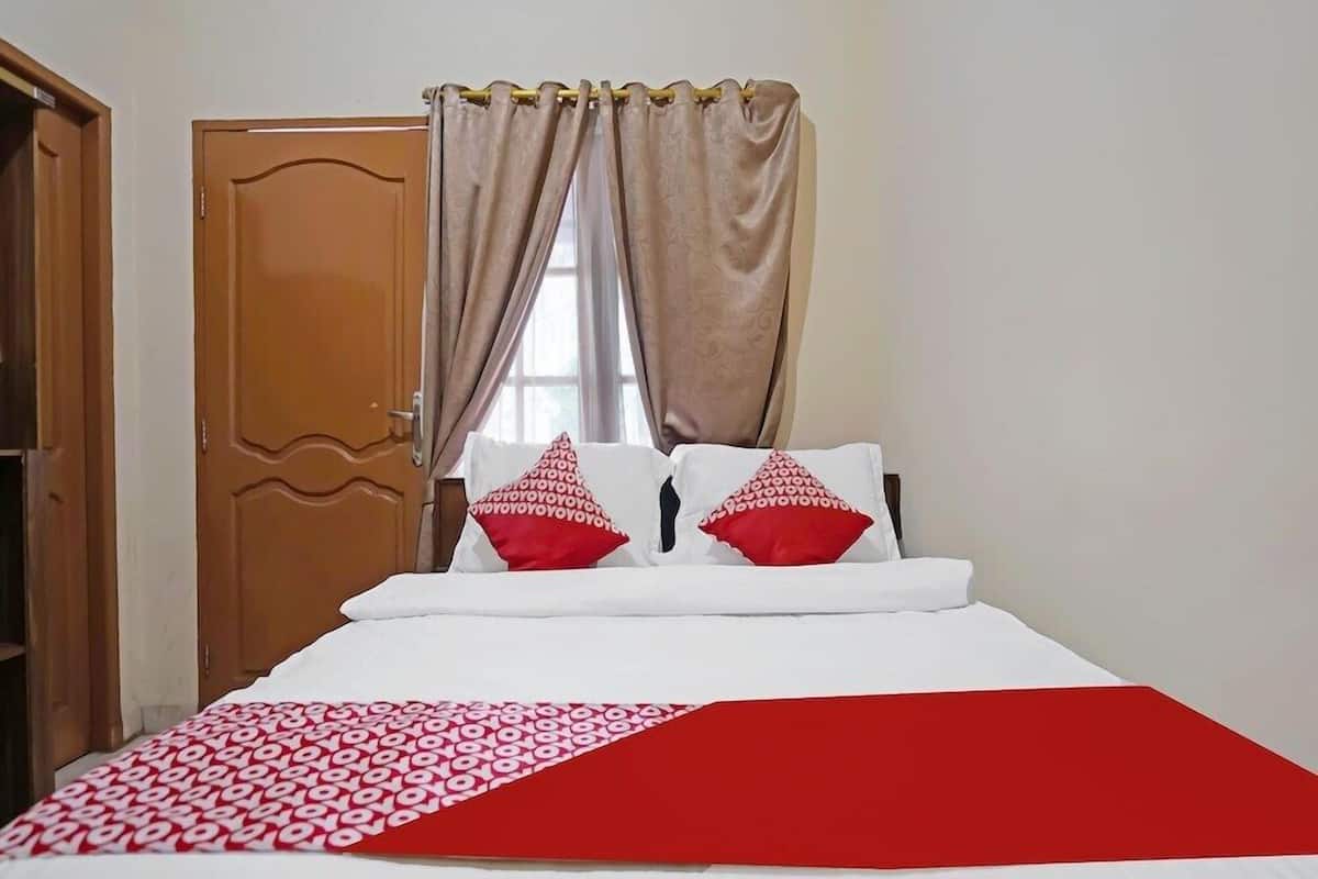 Une chambre à coucher avec un lit, deux oreillers, une couverture rouge et blanche et des rideaux de couleur beige.