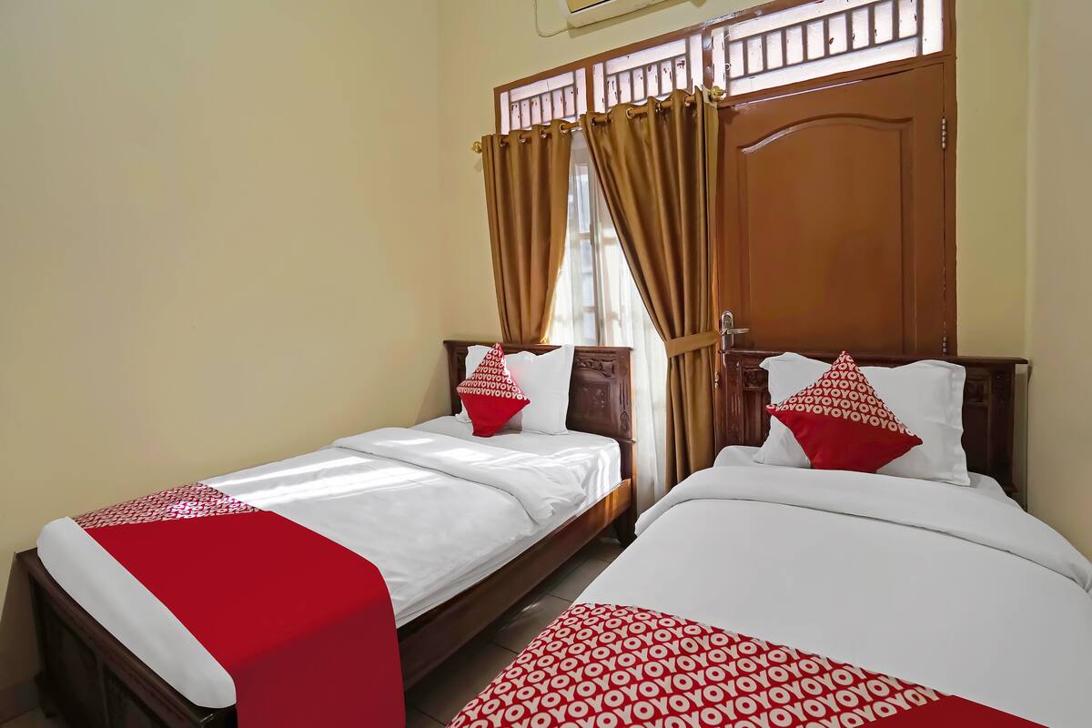 Une chambre d’hôtel avec deux lits, chacun agrémenté de draps blancs et de touches de couleur rouge, une porte en bois et une fenêtre avec des rideaux.