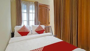 Ein ordentlich gemachtes Bett mit weißer und roter Bettwäsche, einem Holz-Kopfbrett und einem Fenster mit Vorhängen.