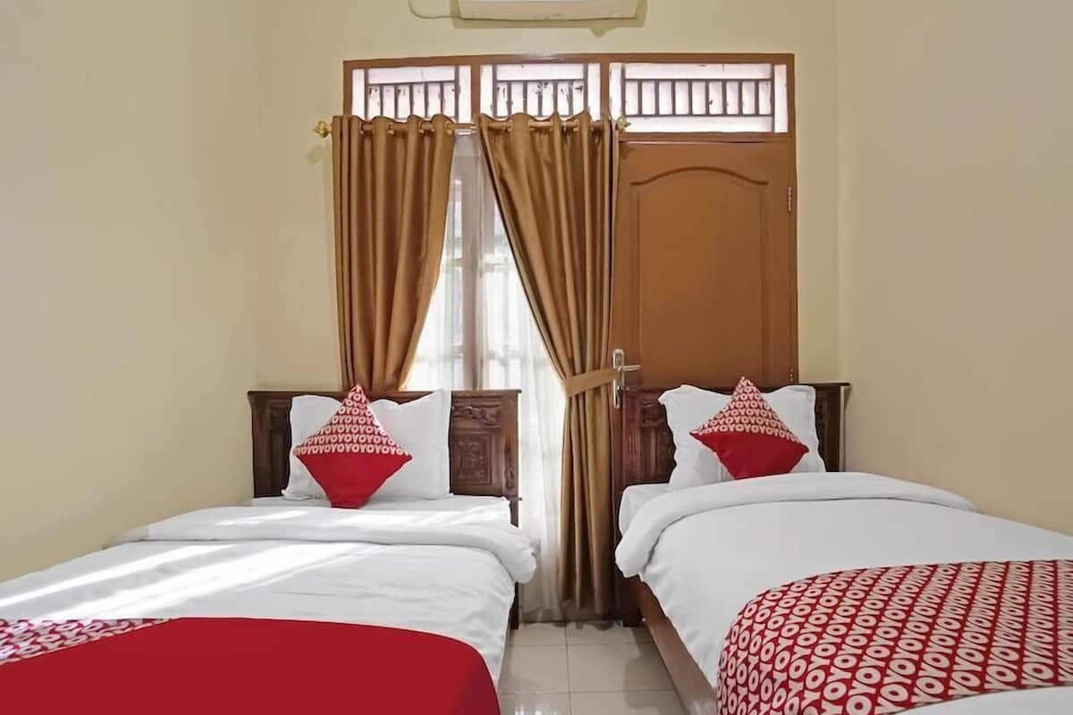 Une chambre d’hôtel avec deux lits simples, une porte en bois, une fenêtre avec des rideaux et un ventilateur de plafond.