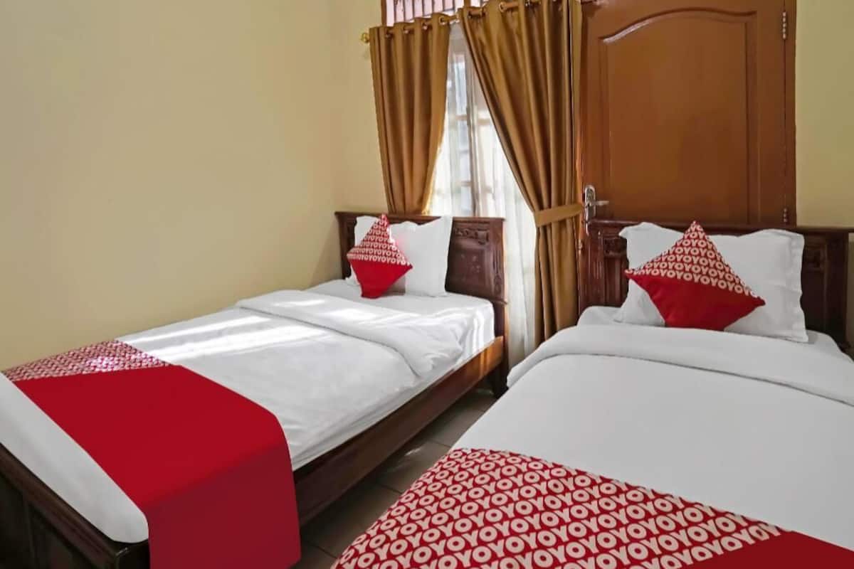 Une chambre d’hôtel avec deux lits, chacun recouvert d’une couvre-lit à motifs rouges et blancs, dotés de têtes de lit en bois et d’une fenêtre avec des rideaux.