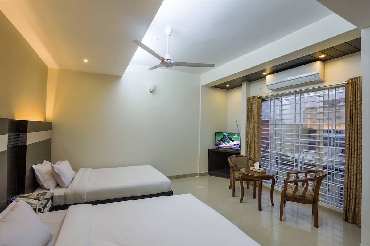 A hotel room with two beds, a ceiling fan, a TV, a window with blinds, and a small table with chairs.