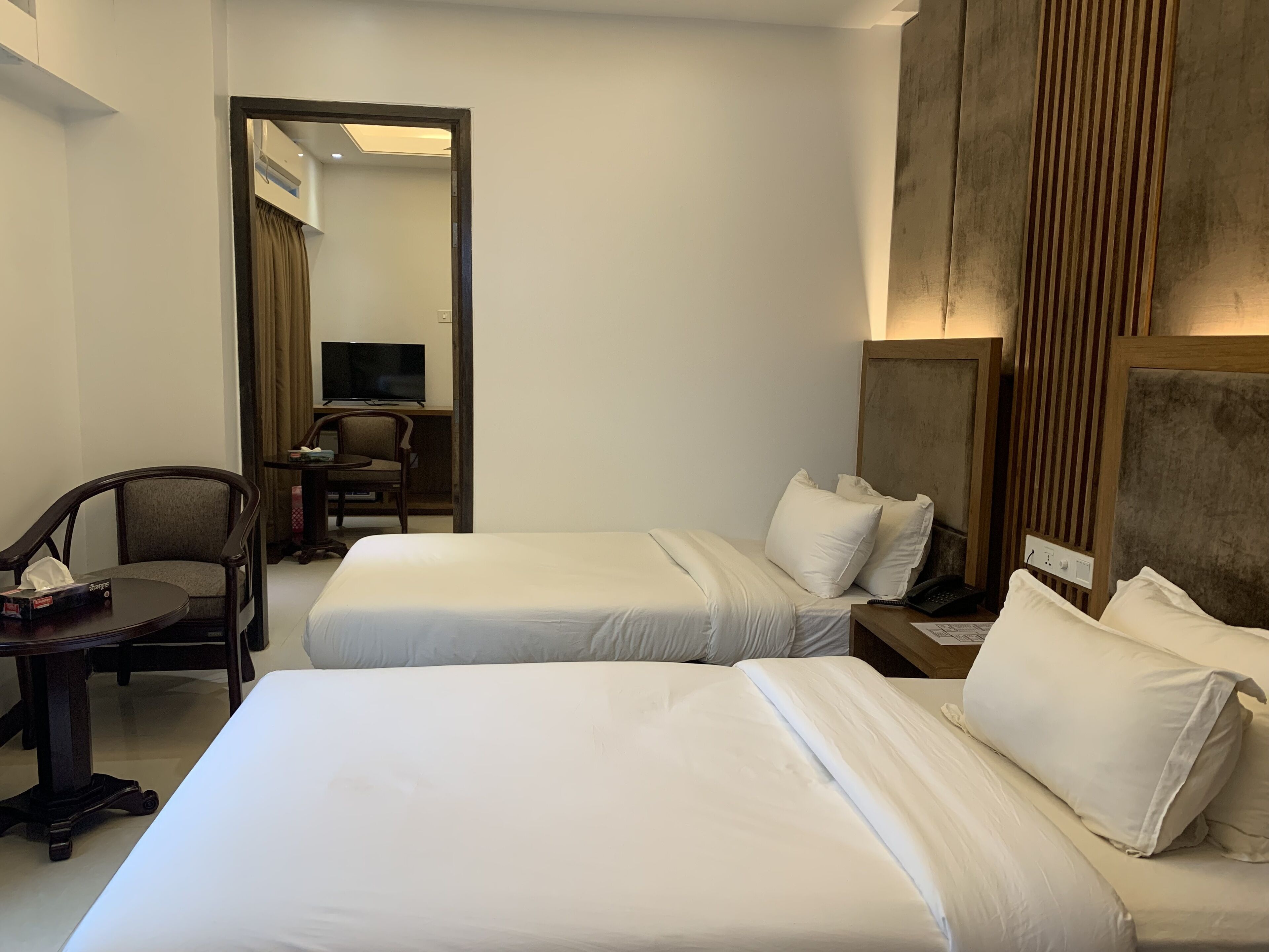 Une chambre d’hôtel avec deux lits, un bureau avec une chaise, une télévision et une tête de lit en bois.