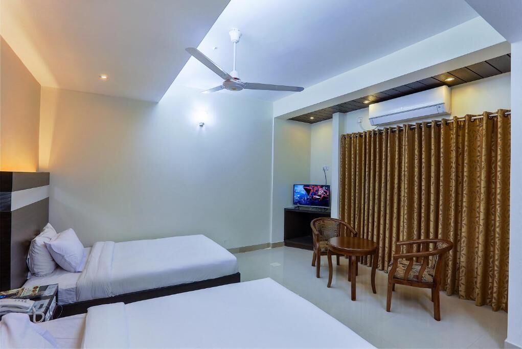 Habitación de hotel con dos camas, ventilador de techo, televisión y zona de comedor con sillas.