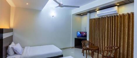 Habitación de hotel con dos camas, ventilador de techo, televisión y zona de comedor con sillas.