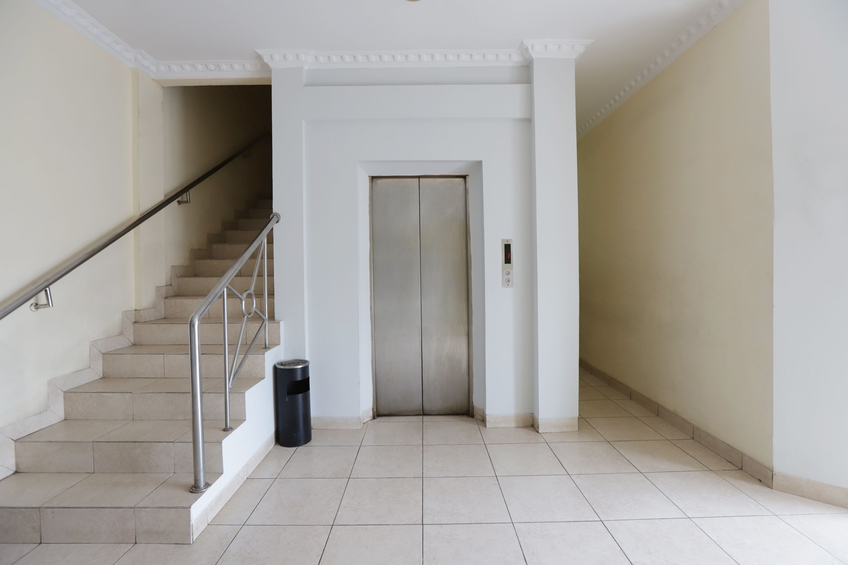 A clean, modern hallway with a staircase, an elevator, and a trash bin.