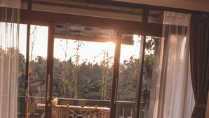 A room with a bed, a wooden bench, and a table with chairs. There is a large window with curtains, and a view of trees and a building outside. - Ubud Paras Villa (Ubud)