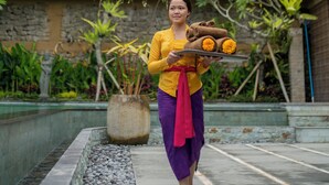 A person carrying a tray with food items, standing in front of a building with a balcony and potted plants. - Ubud Paras Villa (Ubud)