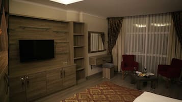 Una sala de estar moderna con un televisor de pantalla plana, armarios de madera, una pequeña mesa con dos sillas y un amplio ventanal con cortinas.