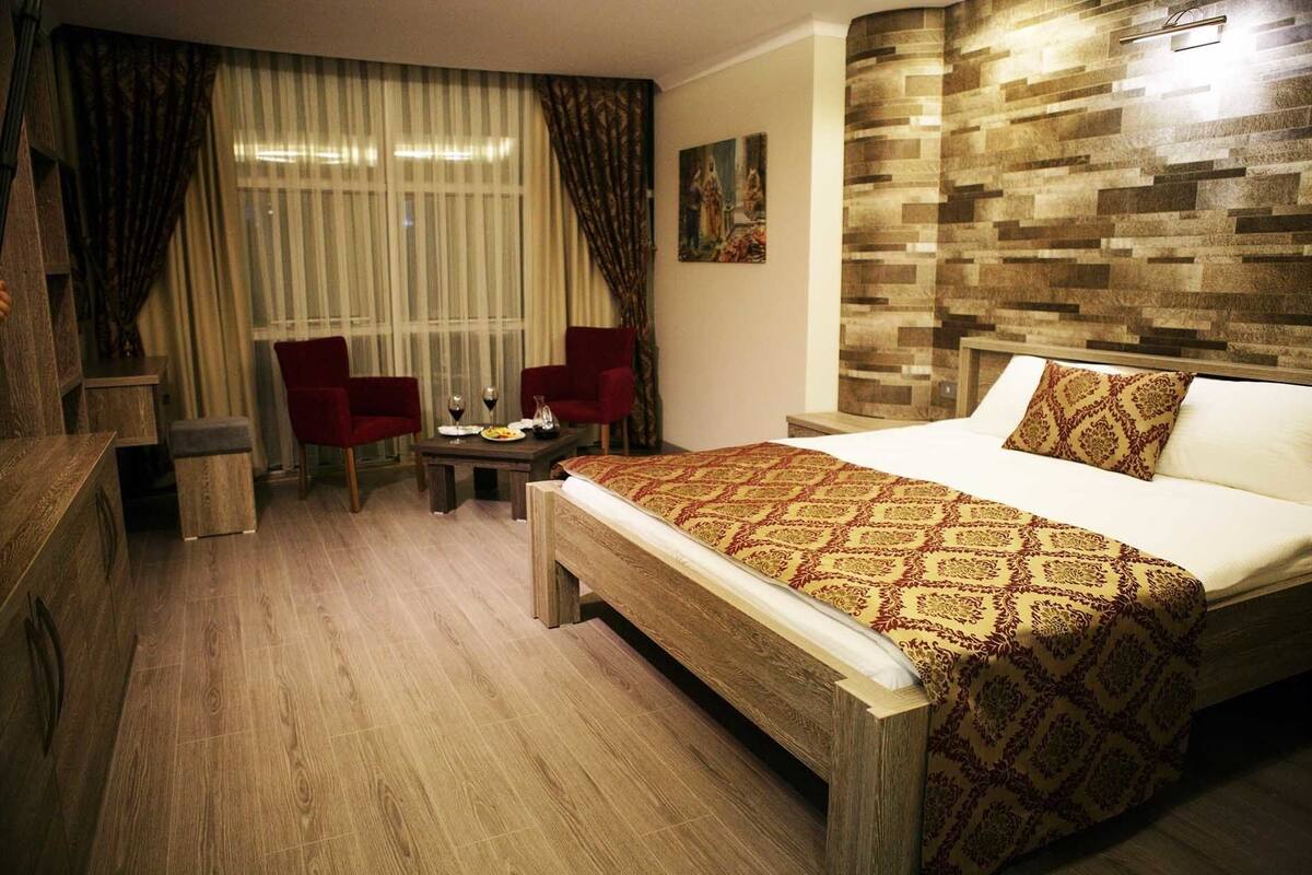 A hotel room with a wooden floor, a bed with a patterned bedspread, two red chairs, a small table with a wine glass, and a painting on the wall.