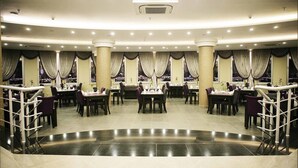 Un restaurant spacieux, doté de nombreuses tables et chaises, d’un espace central ouvert et de grandes fenêtres avec des rideaux.