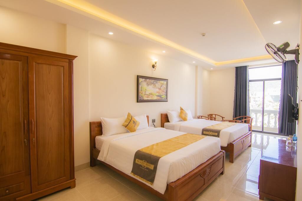 Quarto de hotel com duas camas, um guarda-roupa de madeira, uma escrivaninha e uma janela grande com cortinas.