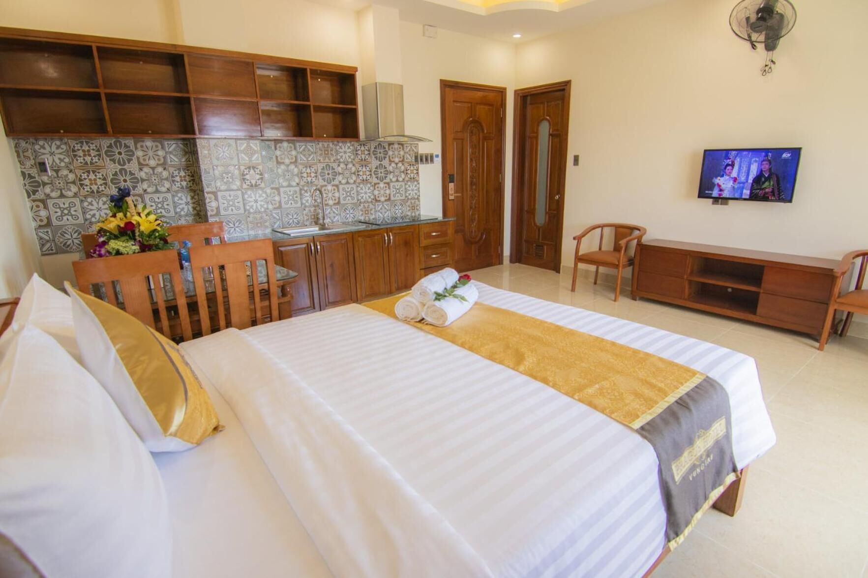 A hotel room with a large bed, a wooden desk, a television, and a kitchenette with patterned tiles.