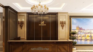 A reception area with a dark wood reception desk, a chandelier, and a large framed view of a cityscape. - Prague Hotel Vung Tau (Ho Chi Minh City)
