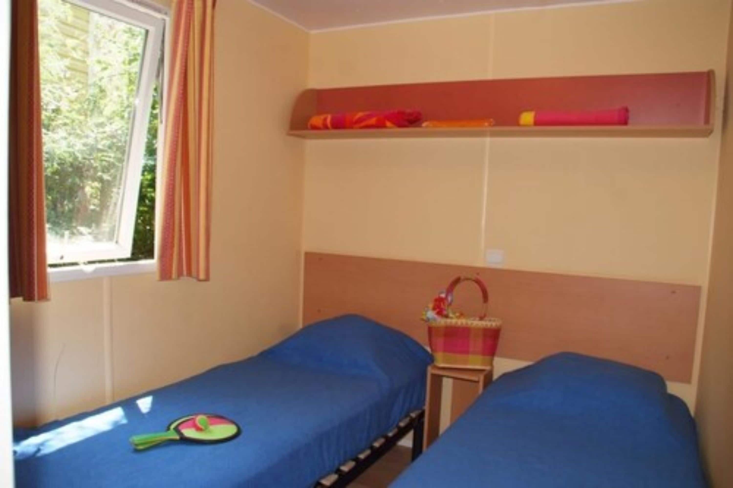 A room with two single beds, a window with curtains, a shelf with towels, and a small table with a basket.