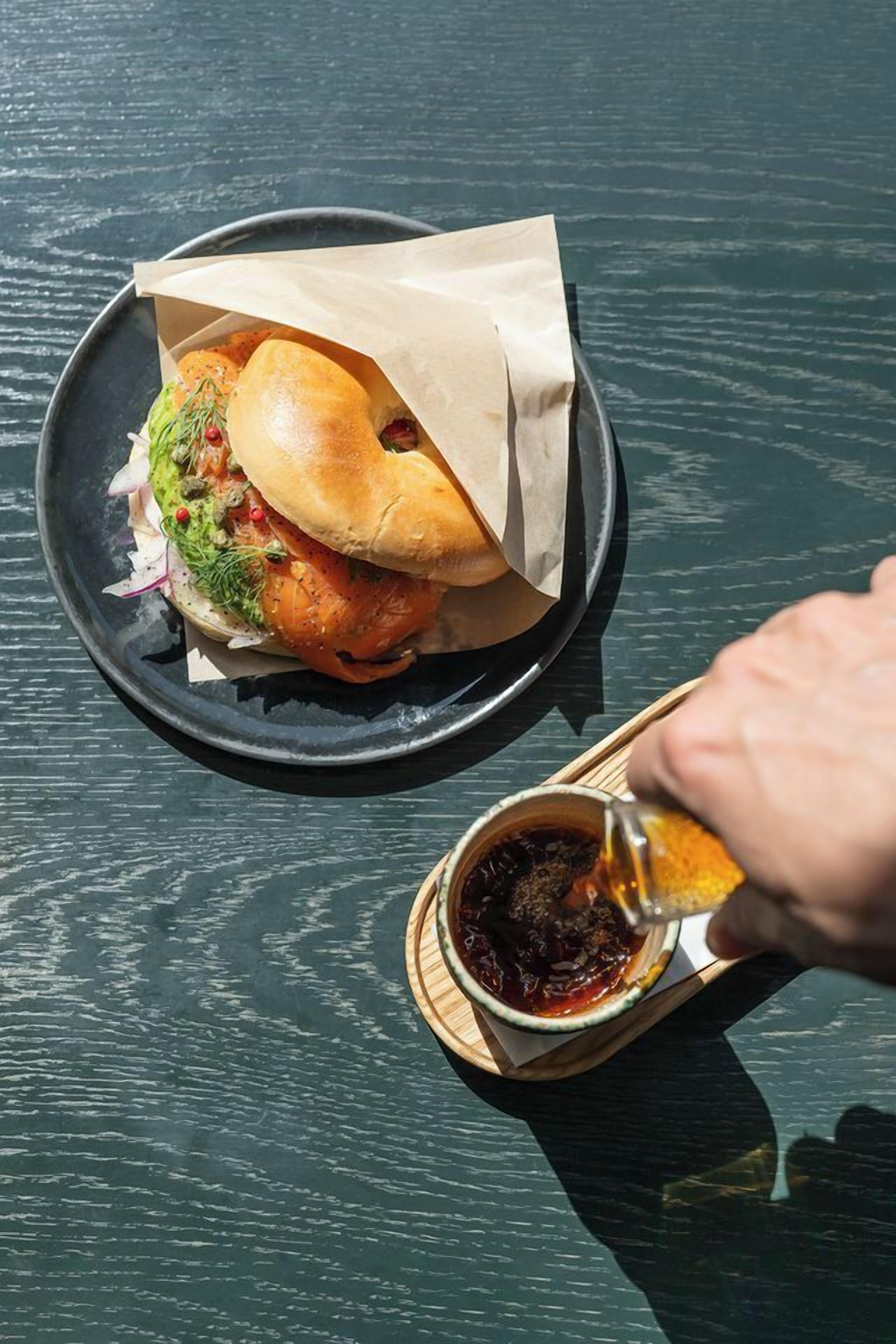 A sandwich on a plate with a paper wrapper, a hand holding a sauce container, and a wooden spoon.