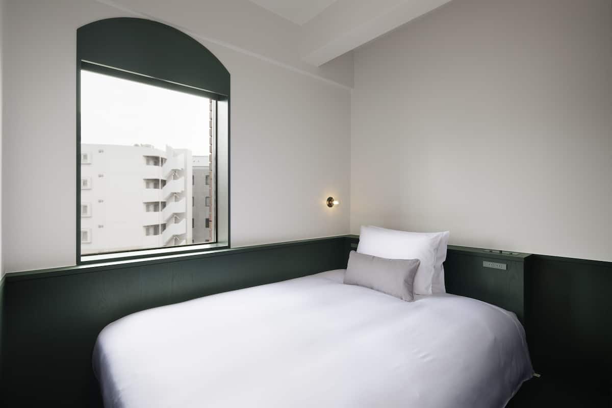 A hotel room with a large bed, a window with a view of buildings, and a wall-mounted light.