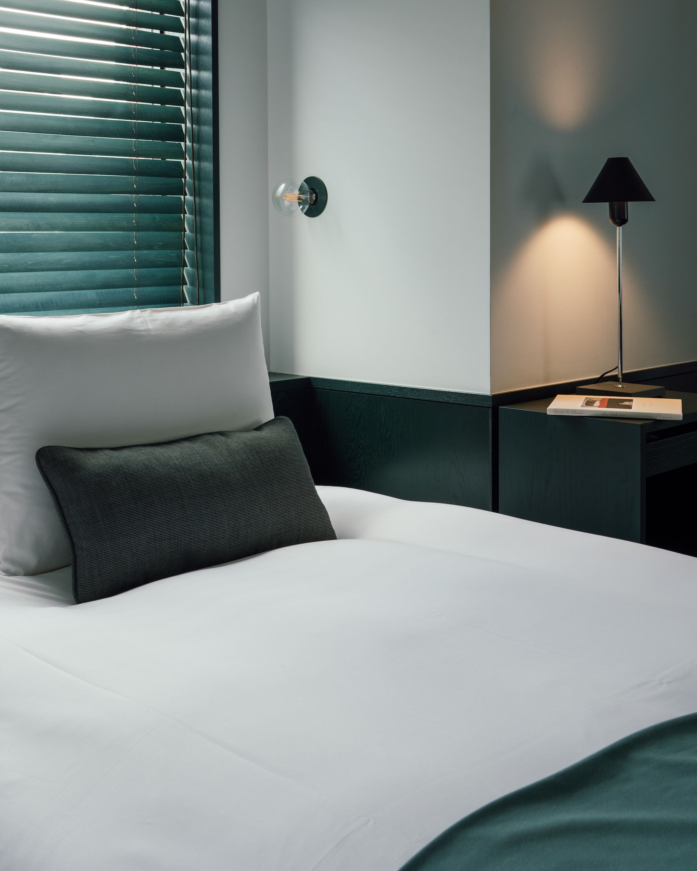 Uma cama bem arrumada com lençóis brancos e travesseiro verde escuro, uma luminária na parede e uma janela com persianas.