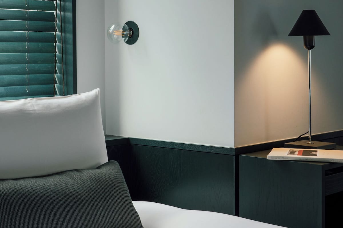 A hotel room with a bed, a nightstand, a lamp, and a wall-mounted light fixture.