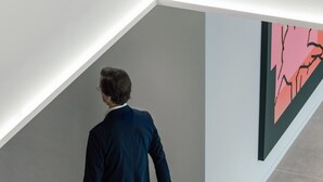 Una persona amb un vestit fosc camina amb una maleta sobre rodes en un passadís modern, amb solera, i una obra d'art minimalista a la paret.