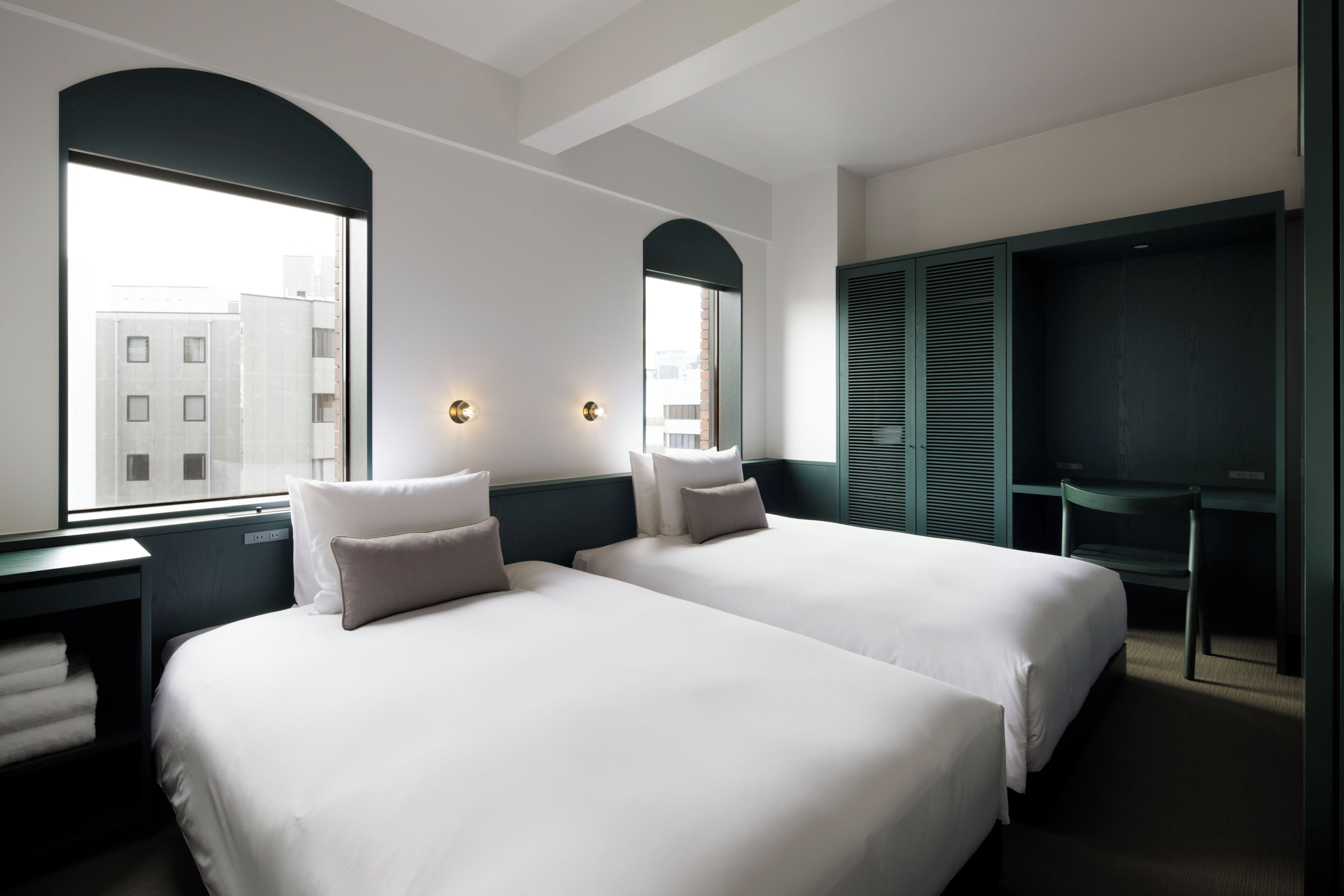 A hotel room with two beds, a desk, a chair, and a large window with a view of buildings.