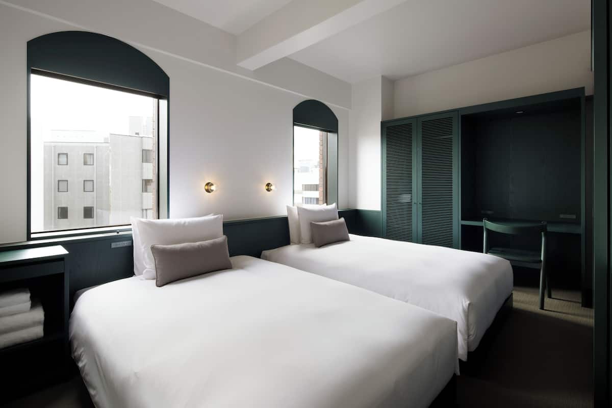A hotel room with two beds, a desk, a chair, and a large window with a view of buildings.