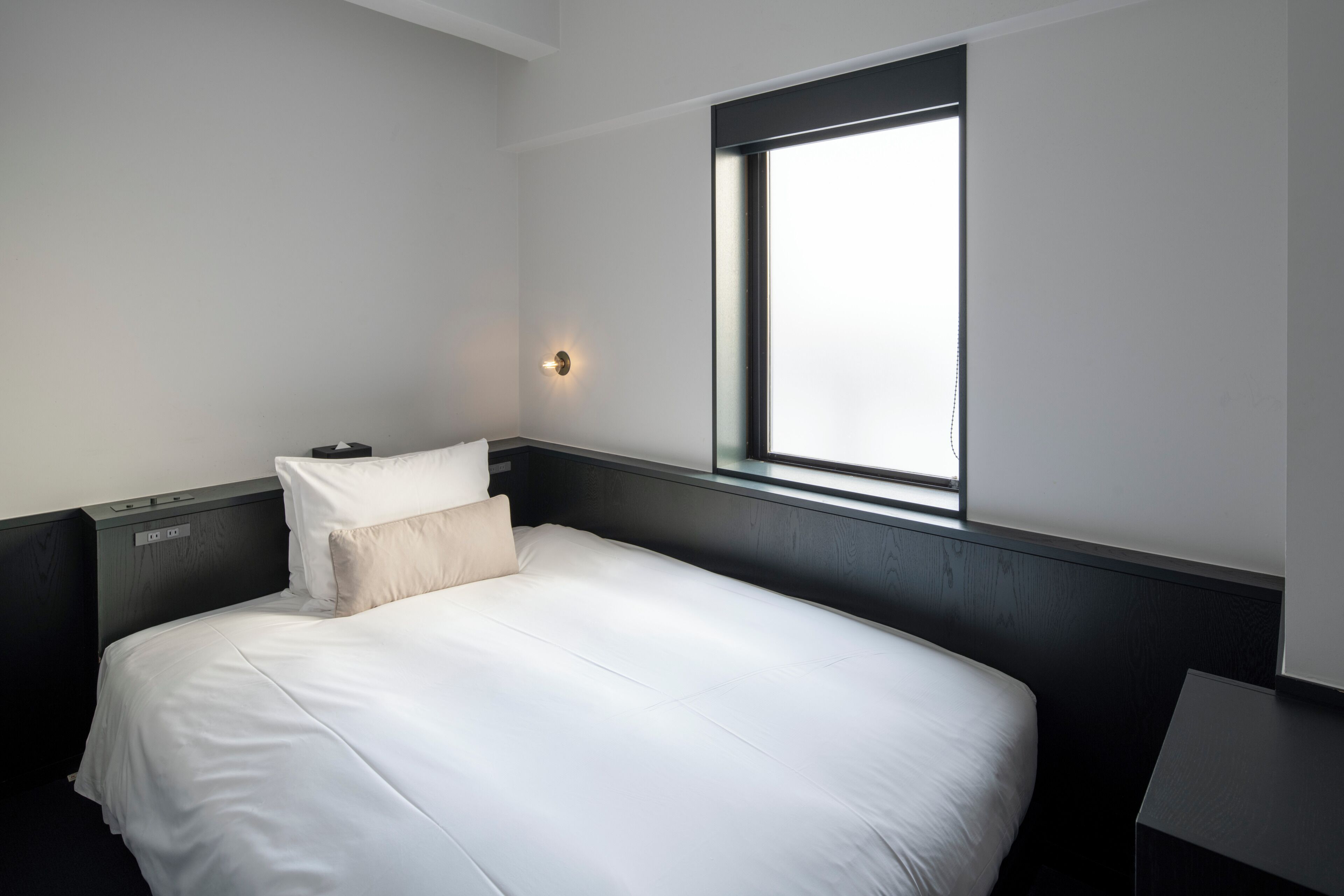 Un lit simple avec une literie blanche, dans une pièce dotée d’une fenêtre, d’une lampe fixée au mur et d’une tête de lit de couleur foncée.