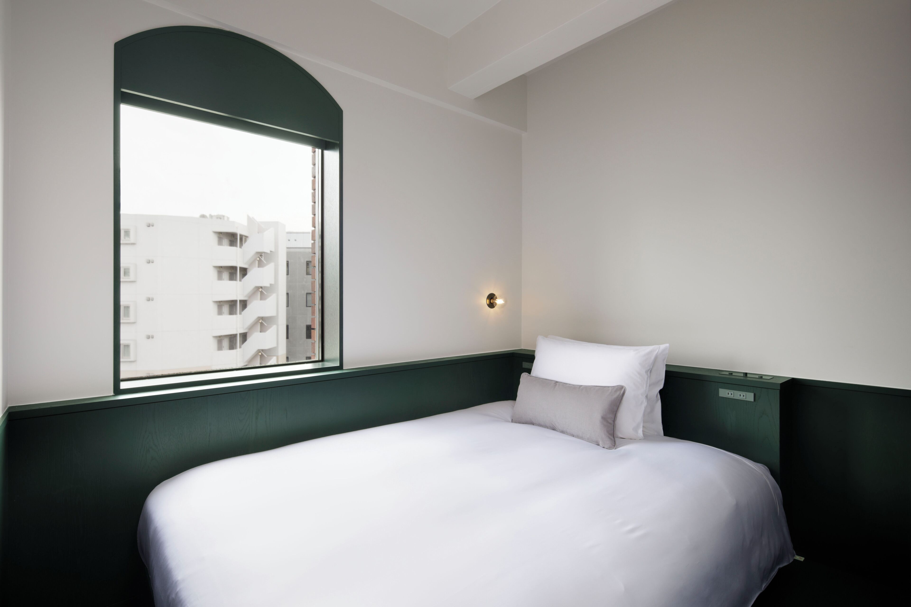 A hotel room with a bed, a window, and a wall-mounted light.