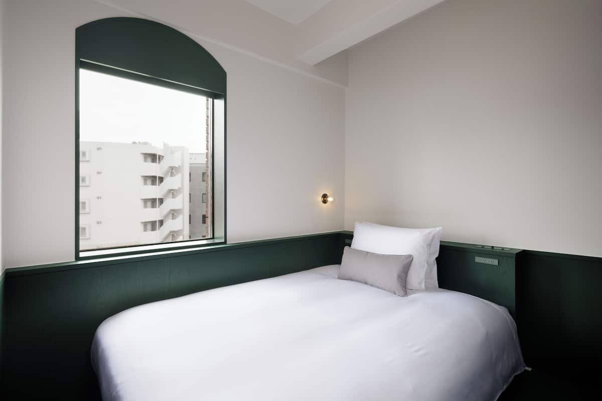 A hotel room with a bed, a window, and a wall-mounted light.
