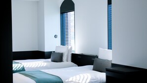 Habitació d'hotel amb dos llits, una tauleta de nit, una làmpada i una finestra amb persianes.