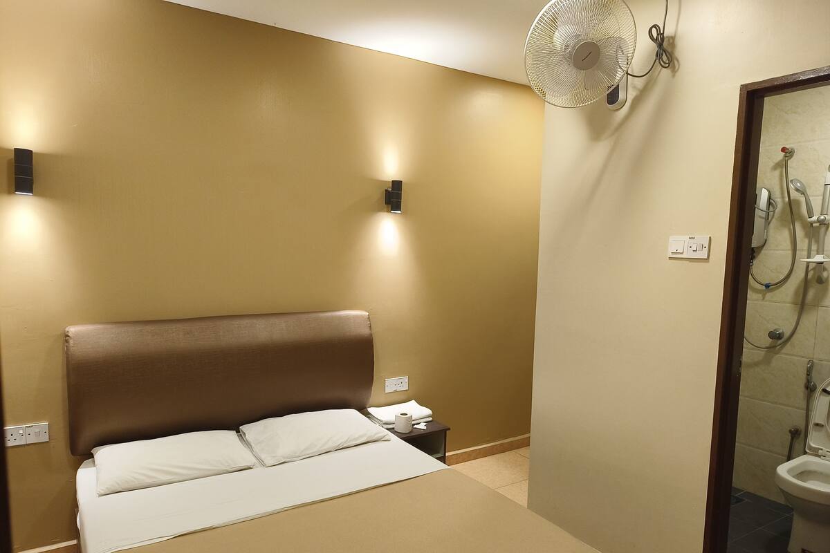 Une chambre d’hôtel avec un lit, une table de chevet, un ventilateur fixé au mur et une vue sur la salle de bain.