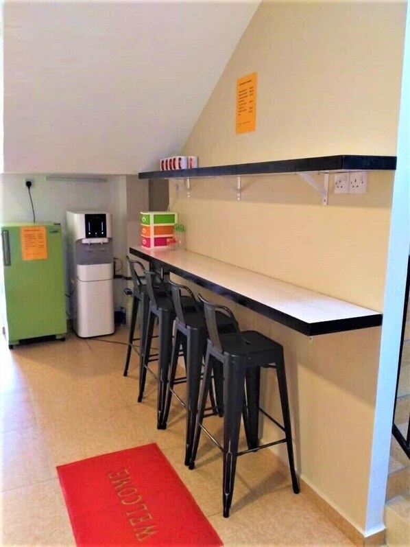 Una kitchenette con refrigerador, dispensador de agua y microondas. Hay cuatro taburetes negros y una alfombra roja con la palabra 'WELCOME' escrita.