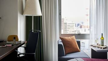 A hotel room with a desk, a chair, a sofa, a lamp, a window with a view of buildings, and a small table with a bottle and glasses.