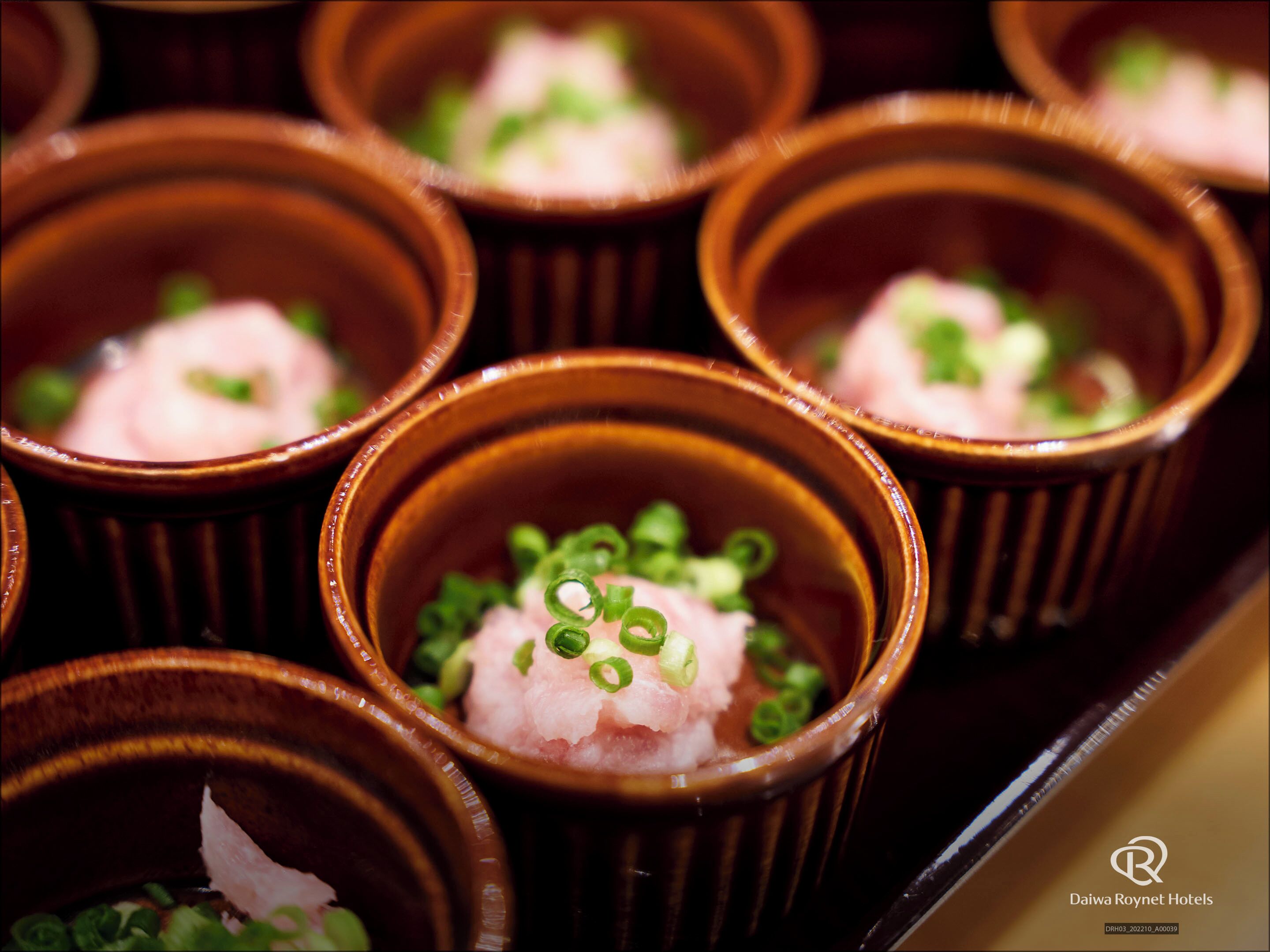 A close-up of several small ceramic dishes containing pink food garnished with green onions, arranged on a tray.