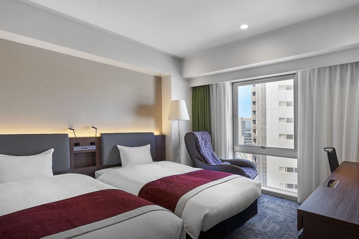Ein Hotelzimmer mit zwei Betten, einem Schreibtisch, einem Stuhl, einer Lampe und Blick auf die Stadt.