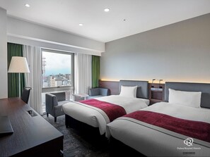 Two beds with white linens and red throw pillows, a desk and chair, and a city view from the window.