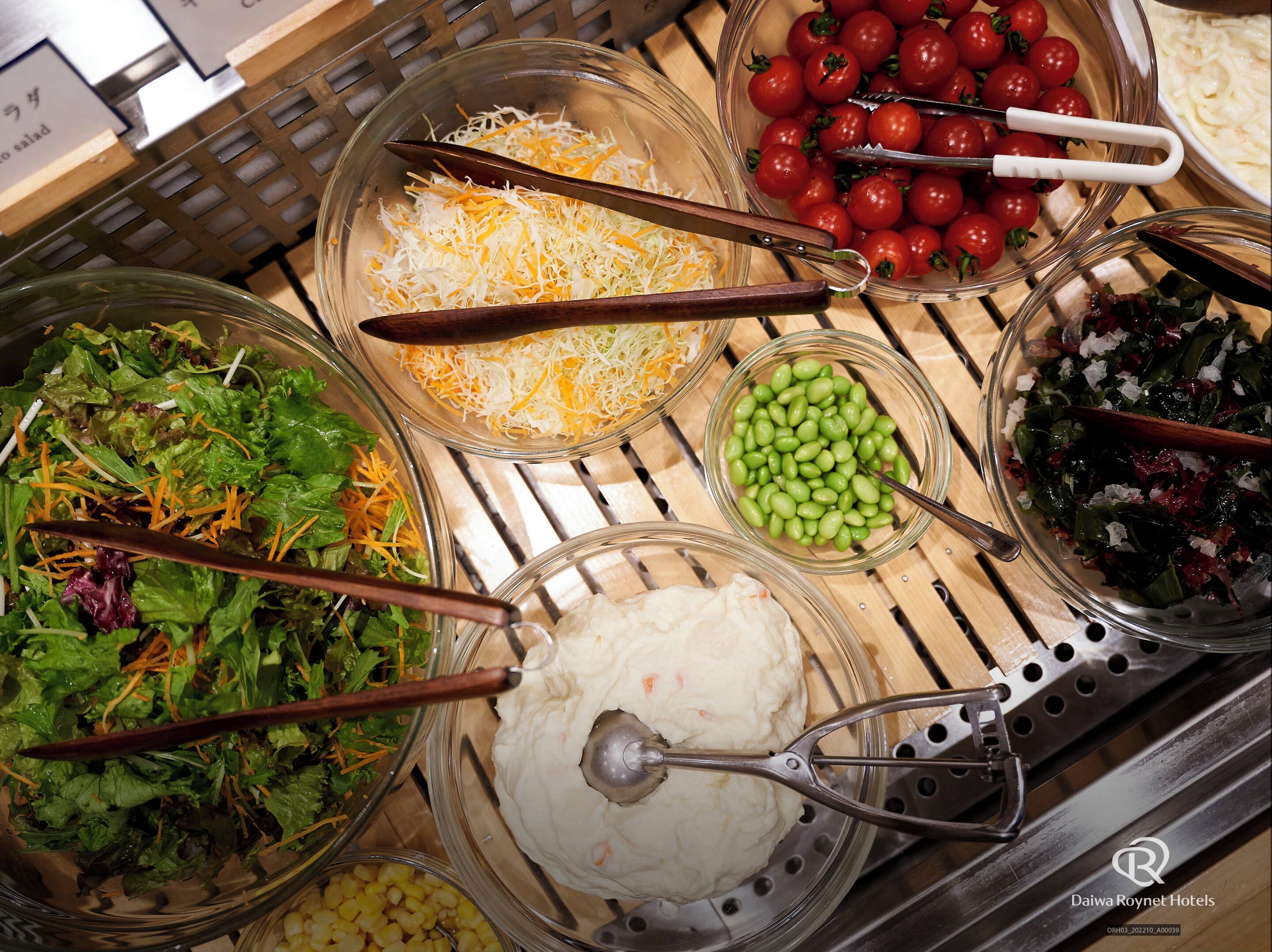 A buffet with various food options including salads, shredded cheese, cherry tomatoes, edamame, and a creamy dip.