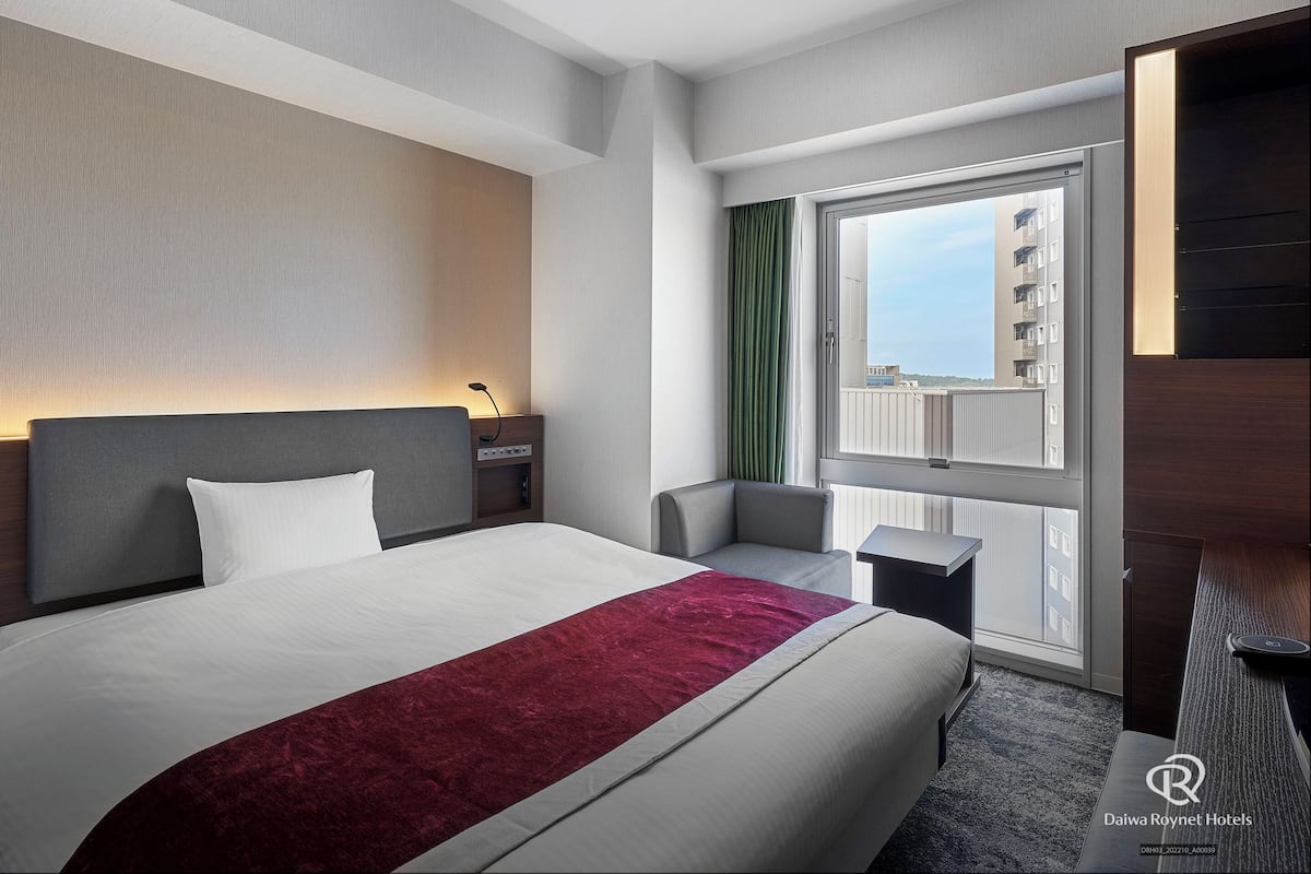 Ein Hotelzimmer mit Bett, Nachttisch, Stuhl und einem großen Fenster mit Blick auf Gebäude.
