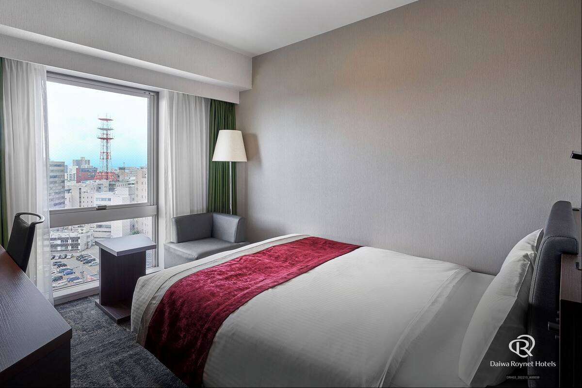 Ein Hotelzimmer mit einem großen Bett, einem Schreibtisch, einem Sessel und einer Lampe, mit Blick auf die Stadt durch ein großes Fenster.