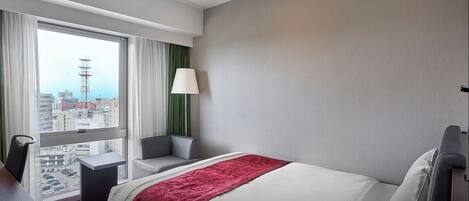 A hotel room with a large bed, a desk, a chair, a lamp, and a view of the city through a large window.