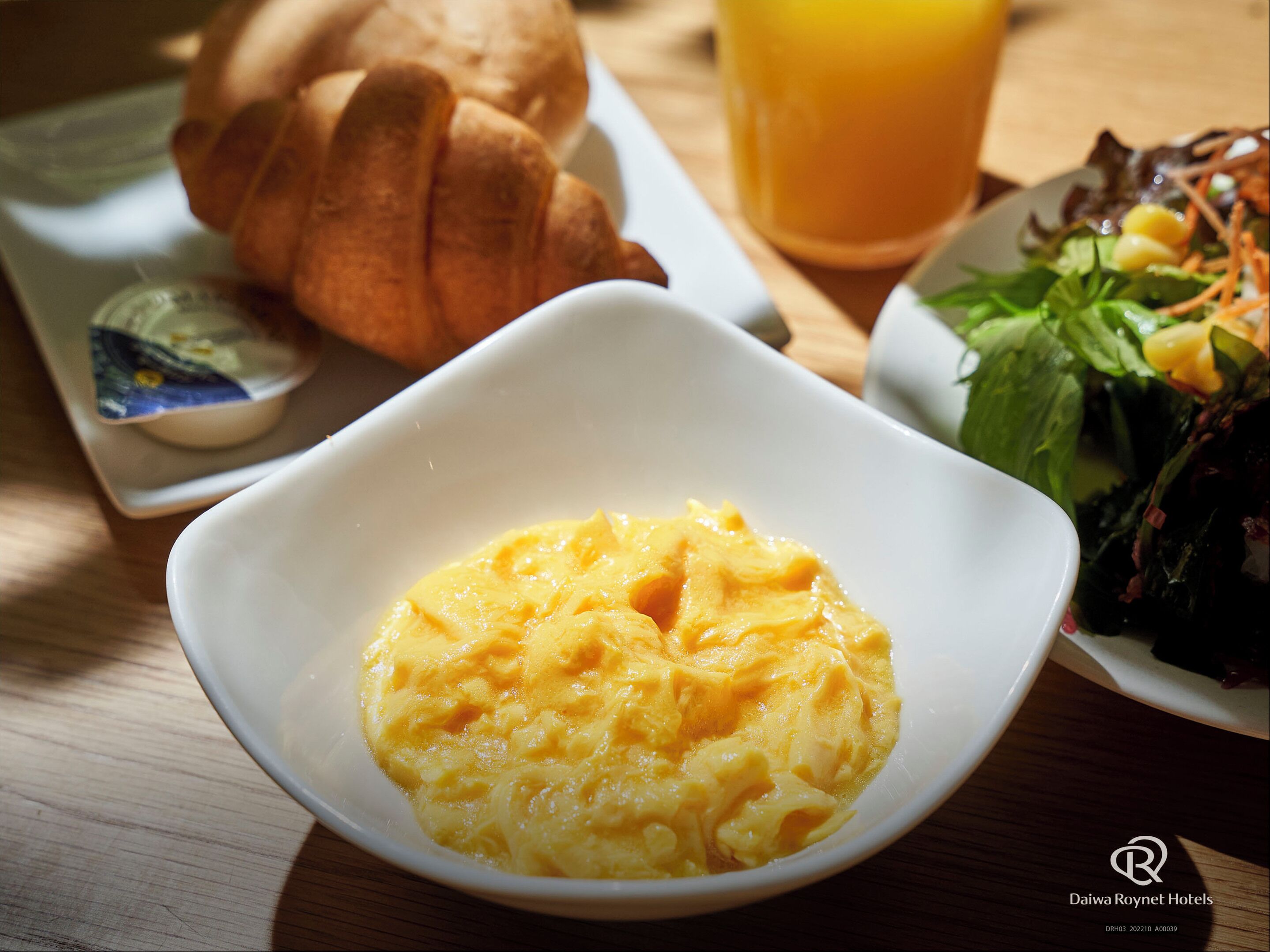 Une table en bois dressée pour le petit-déjeuner, avec des œufs brouillés, un croissant, un verre de jus d’orange et une petite salade.
