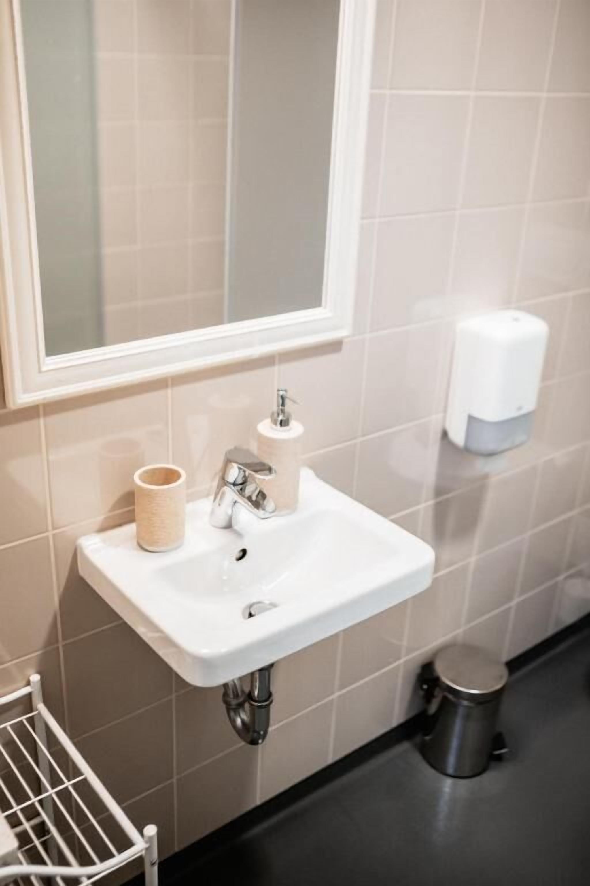 Un baño con un lavamanos blanco, un espejo, un dispensador de toallas y un cubo de basura.