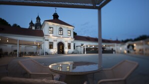 A covered outdoor seating area with wicker chairs and a glass table, in front of a white building with a red roof and multiple domes, during twilight. - Hotel Monte Pacis (Kaunas)