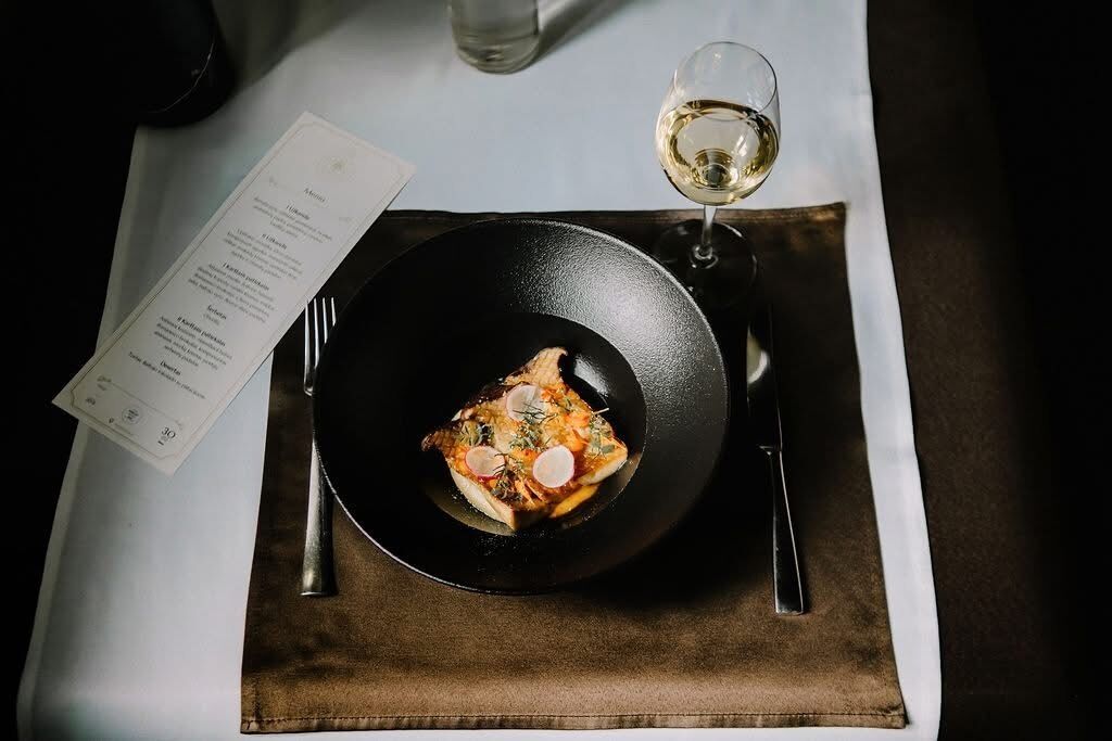 A black bowl of food on a table with a menu and a glass of white wine.