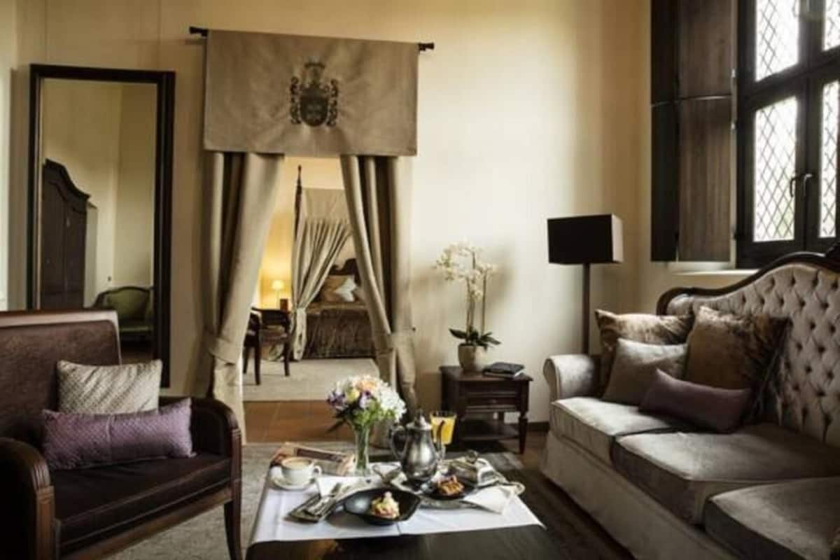 A room with a dining table set for breakfast, a sofa, a lamp, and a large window with curtains.