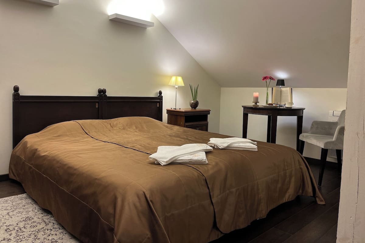 A hotel room with a large bed, a desk with a lamp, a chair, and a small table with a candle and flowers.