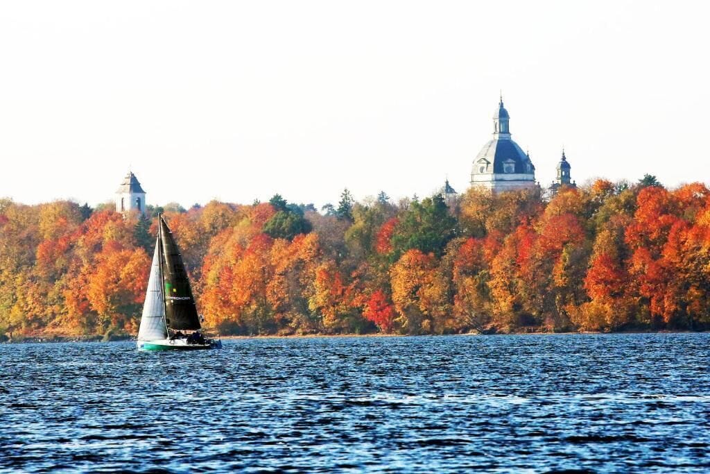 A sailboat on a lake with autumn foliage and a building in the background.